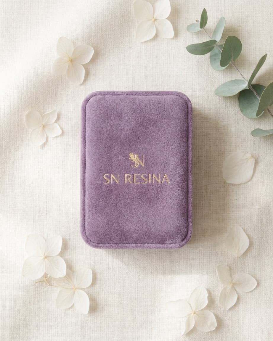 Caixinha de veludo lilás SN Resina com monograma dourado, rodeada de flores brancas e folhas de eucalipto